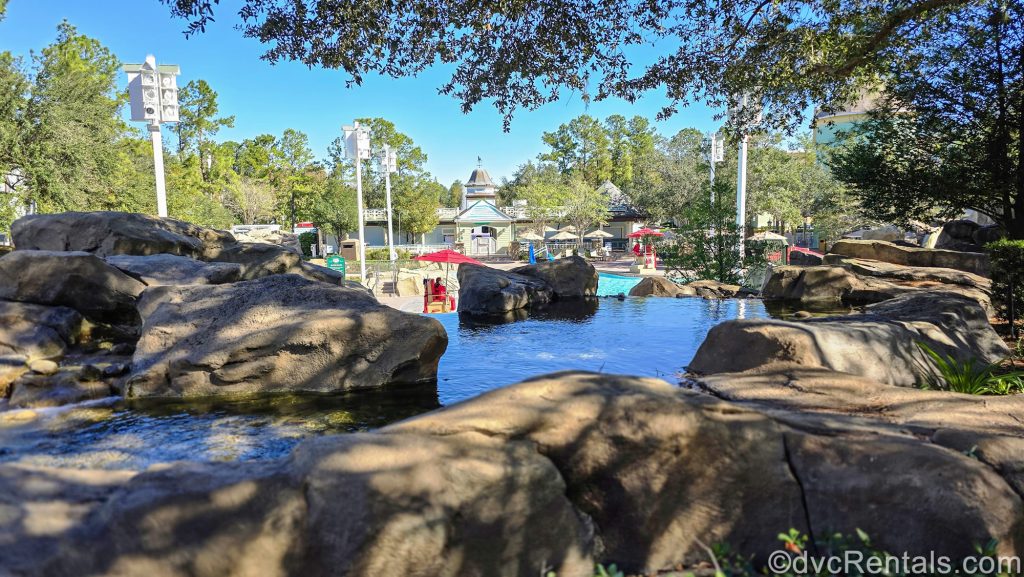 The High Rock Spring pool at Disney’s Saratoga Springs Resort & Spa is seen from an upper rock-based water feature while the pool and surrounding landscaping are shown below under a sunny sky.