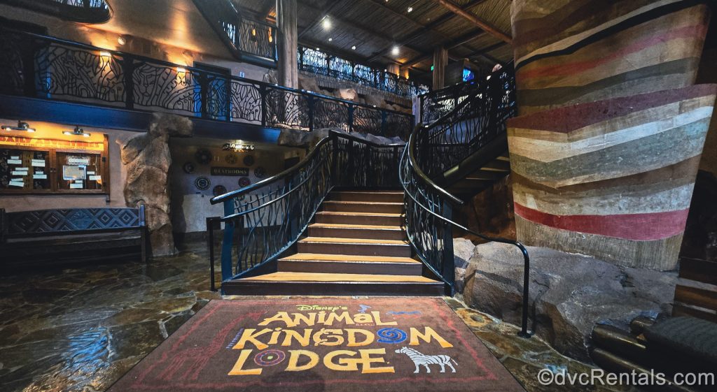 A large rug reading “Disney’s Animal Kingdom Lodge” in yellow writing and decorated with multicolored animals is seen at the base of a stairway near the resort lobby.