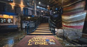 A large rug reading “Disney’s Animal Kingdom Lodge” in yellow writing and decorated with multicolored animals is seen at the base of a stairway near the resort lobby.