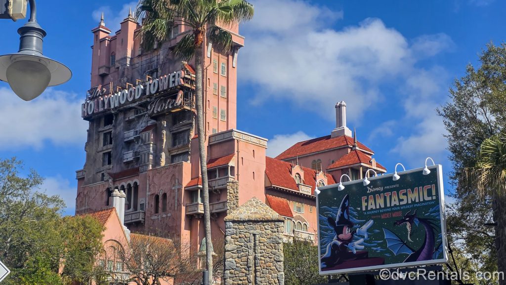 The exterior of the Twilight Zone Tower of Terror is seen under a blue sky next to a billboard for Fantasmic! at Disney’s Hollywood Studios Theme Park.