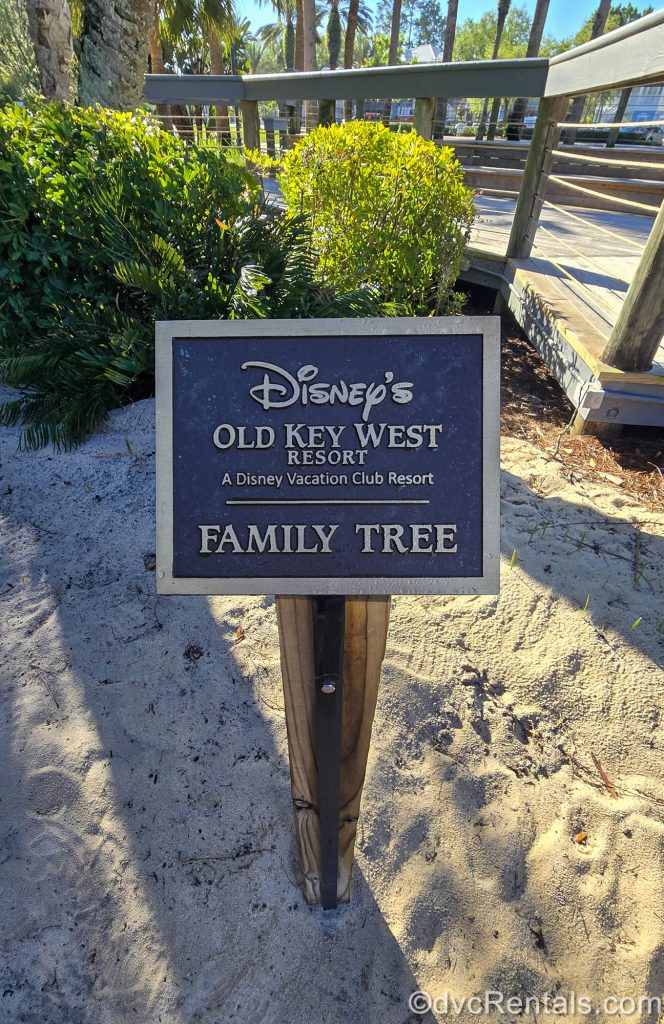 A sign reading “Disney’s Old Key West Resort, a Disney Vacation Club Resort, Family Tree” is seen in the sand with landscaping and resort grounds in the background.