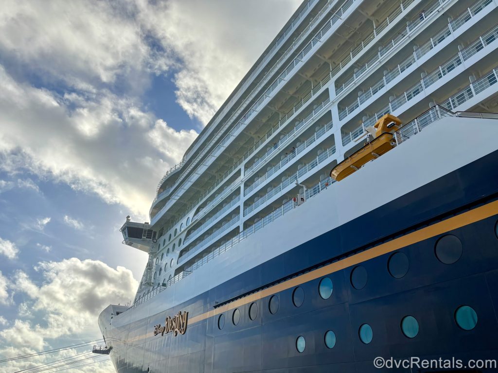 The white and navy-blue exterior of the Disney Destiny cruise ship is seen from the ground up under a sunny blue sky. The ship’s name is written in yellow on the side.