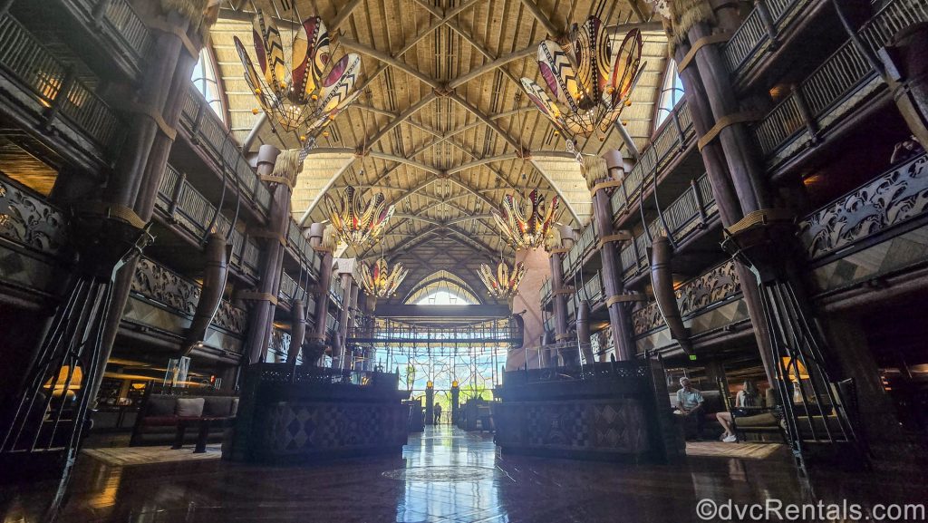 The grand lobby of Disney’s Animal Kingdom Villas Jambo House. The lobby is decorated with warm tones and dark wood, and there are displays of African art and artifacts for guests to enjoy around the seating areas.