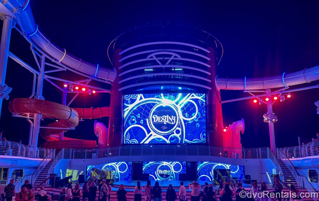 Guests gather in front of the Disney Destiny’s funnel screen and stage under a night sky as the ship’s logo is displayed.
