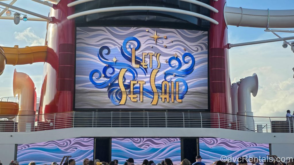 The words “Let’s Set Sail” are displayed on the Disney Destiny’s funnel screen during the Sail Away Deck Party under a sunny sky as guests gather below.