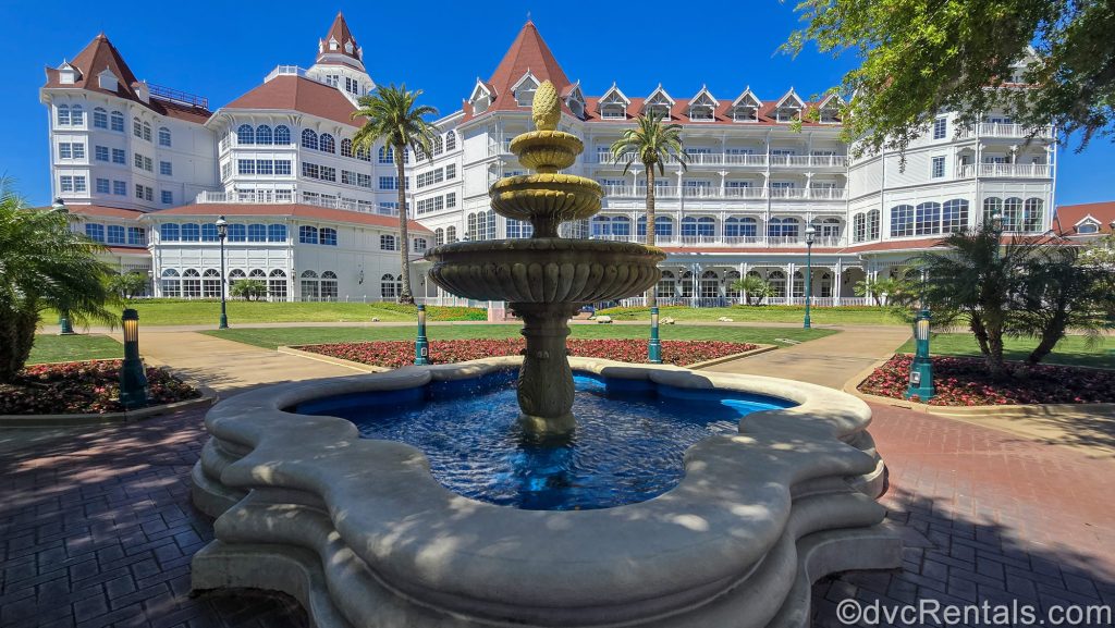 Water lightly trickles from a stone fountain outside Disney’s Grand Floridian Resort & Spa. The iconic white buildings with red roofing are seen in the background across a green lawn landscaped with red flowers.