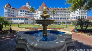 Water lightly trickles from a stone fountain outside Disney’s Grand Floridian Resort & Spa. The iconic white buildings with red roofing are seen in the background across a green lawn landscaped with red flowers.