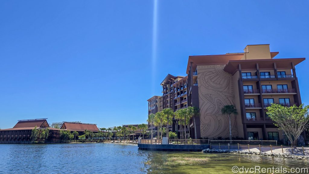 The brown exterior buildings and green palm trees of Disney’s Polynesian Villas & Bungalows and the Island Tower are seen under a bright blue sky along the shores of the Seven Seas Lagoon.