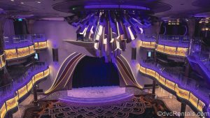 The Grand Hall on the Disney Destiny is seen from an upper deck. The space is lit up with purple and gold effects.