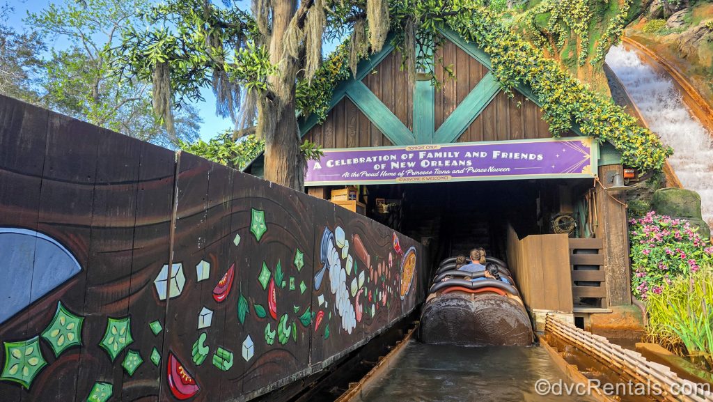 Guests ride Tiana’s Bayour Adventure at Magic Kingdom. The logs float along the water under a purple sign reading “A celebration of family and friends of New Orleans” in white lettering.
