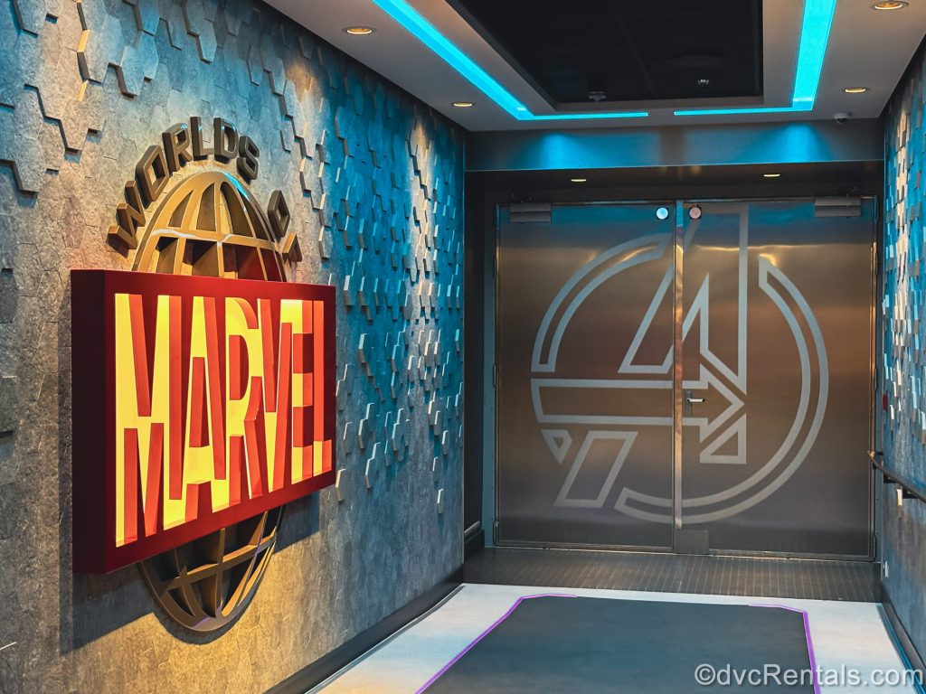 The entry to the Worlds of Marvel dining room on the Disney Destiny is seen. On the left wall the name of the restaurant is displayed in red and gray across a large silver globe design, and the “A” design of the Avengers logo is displayed on the steel entry door.