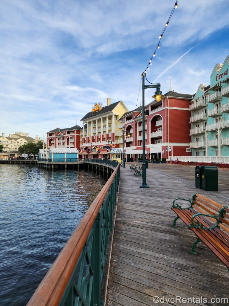 The colorful buildings of Disney’s BoardWalk Villas are seen under a blue sky as they line the waters of Crescent Lake along the wooden boardwalk.