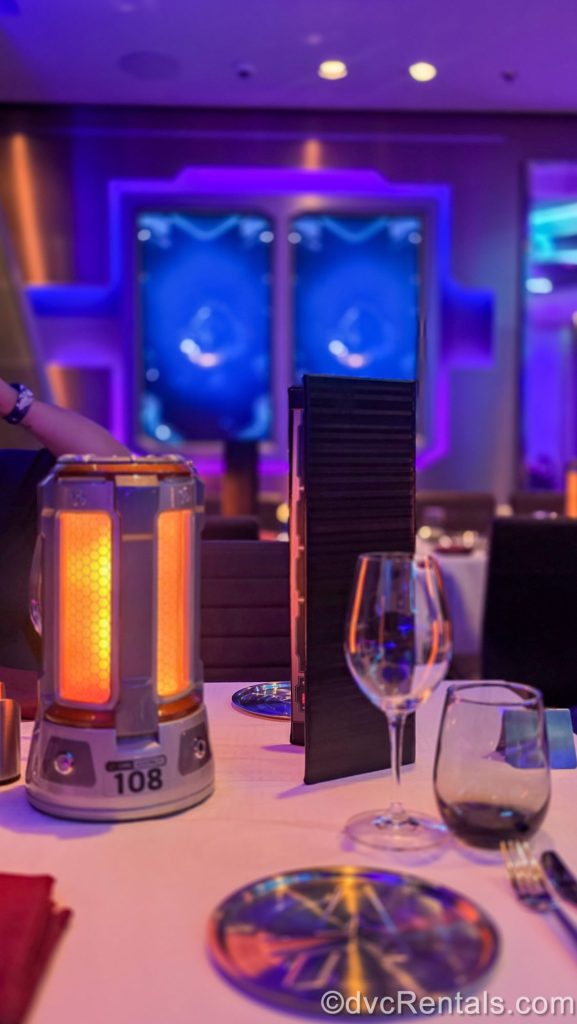 The place settings of the tables and décor in the Worlds of Marvel restaurant are seen as the dining room is lit with purple, blue, and orange lighting.