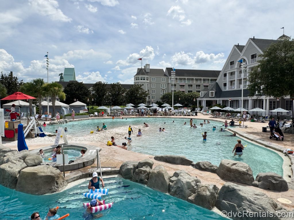 Guests enjoy the water and sand features of the Stormalong Bay pool at Disney’s Beach Club Villas under a sunny, blue sky.