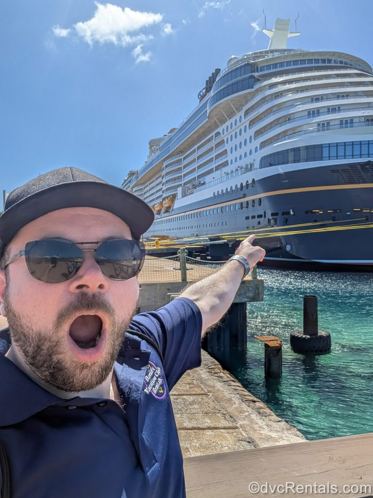 David’s Vacation Club Rentals team member Zac poses with a surprised expression in front of the Disney Destiny cruise ship under a sunny blue sky.