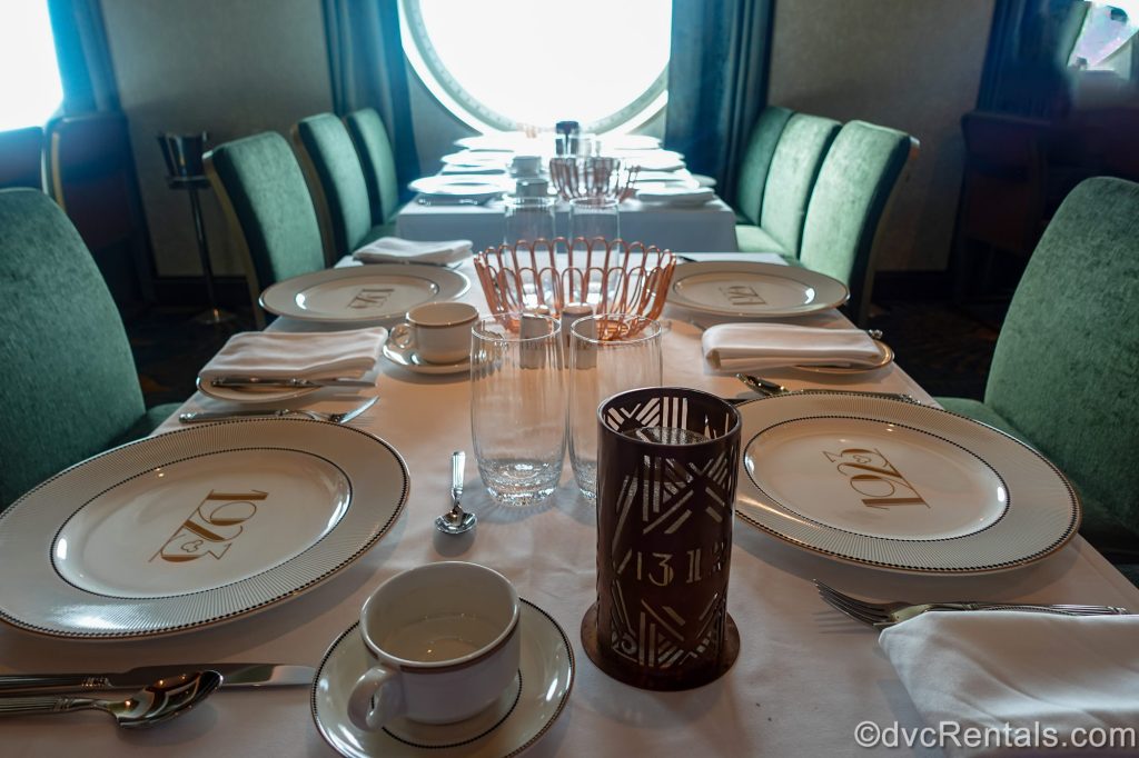 Place settings featuring the dining room’s name, 1923, are seen set on a table with green chairs and a white tablecloth.