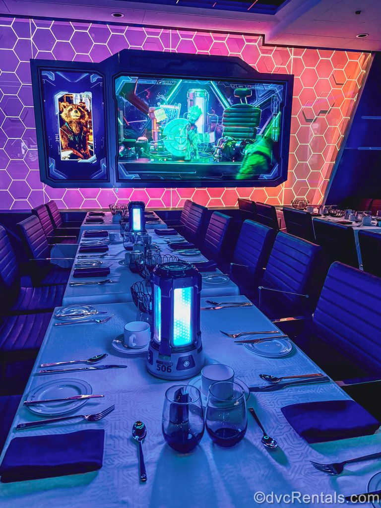 Tables are set for guests to enjoy dinner in the Worlds of Marvel dining room on the Disney Destiny. Behind the tables are TV screens playing Marvel trivia with characters from the Guardians of the Galaxy franchise.