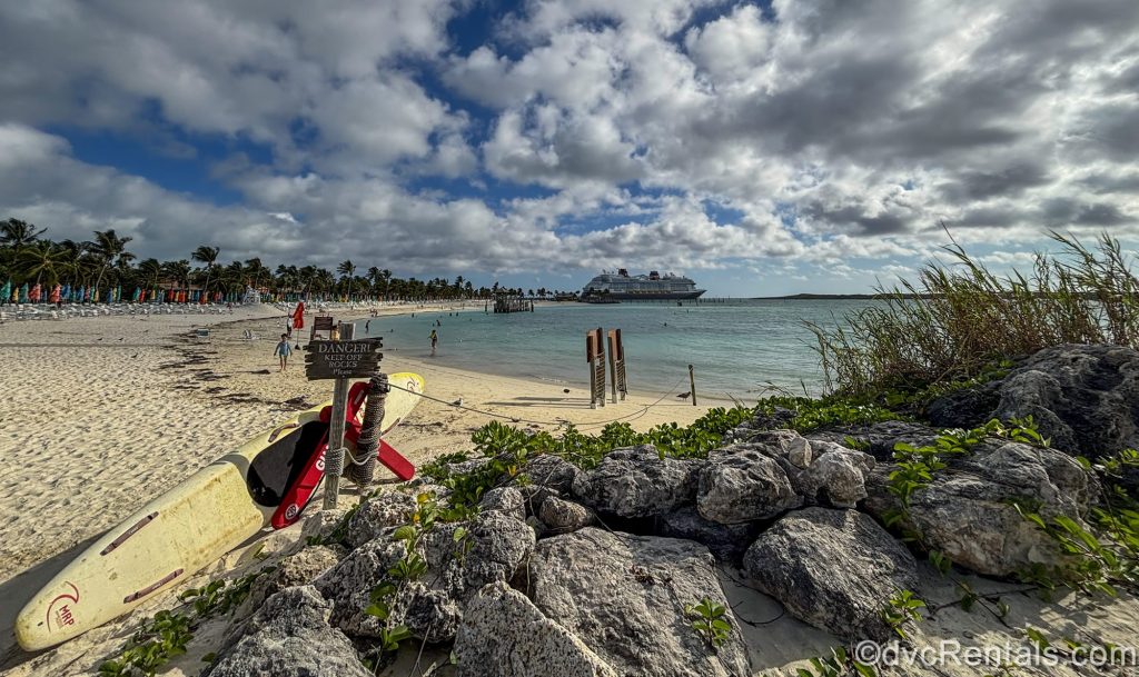 The Disney Destiny is seen in the distance from across the beach and water at Disney Castaway Cay under a blue sky full of fluffy clouds.