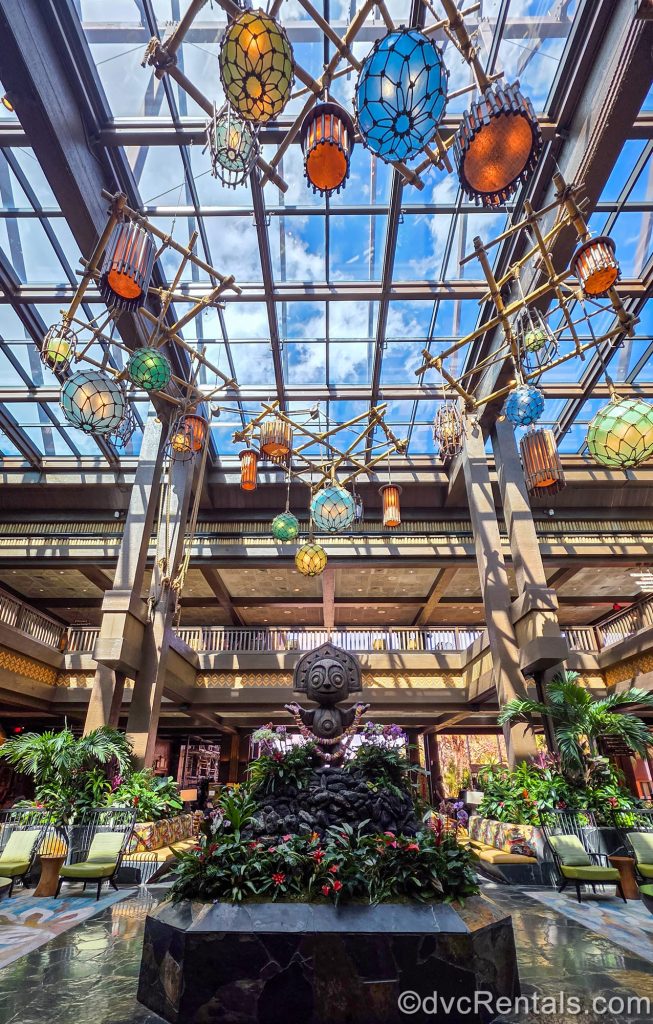 A Tiki statue surrounded by green foliage and flowers is seen in the center of the Great Ceremonial House lobby. Multicolored lanterns hang from the glass ceiling and there are seating areas in the background.
