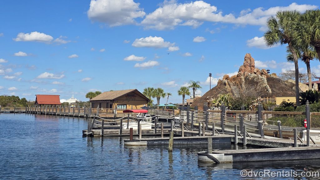 Marina docks at Disney’s Polynesian Villas & Bungalows are seen under a bright sunny sky. The resorts Lava Pool area can be seen to the right and the spires of Space Mountain at Magic Kingdom are seen across the water in the distance.