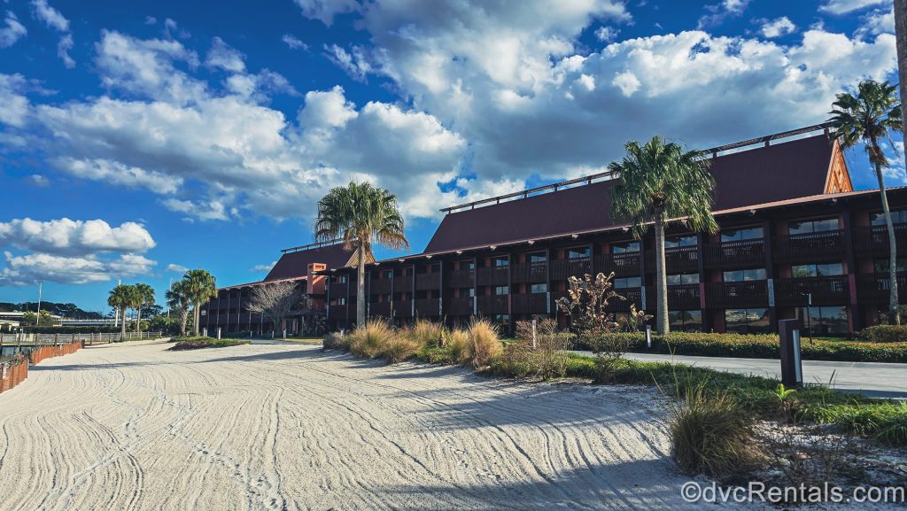 The dark brown exterior of Disney’s Polynesian Villas & Bungalows is seen under a blue sky, with white sand and palm trees lining the Seven Seas Lagoon in front of the buildings.