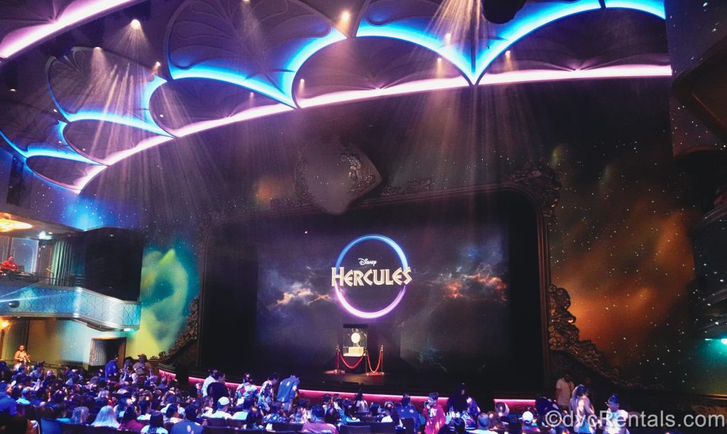 Audience members take their seats in the theater ahead of a performance of Hercules on the Disney Destiny. The show’s logo and a coin prop are displayed on stage.