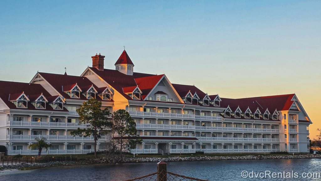 The white exterior and red roof of Disney’s Grand Floridian Resort & Spa is seen during golden hour under a hazy blue and yellow sky.