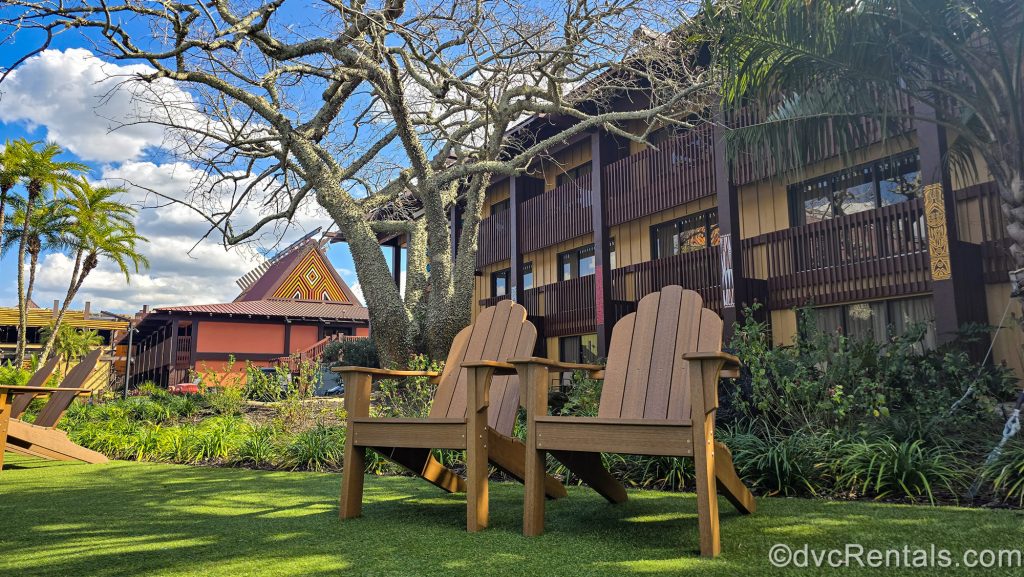 Wooden deck chairs are seen on the grass outside the buildings of Disney’s Polynesian Villas & Bungalows under a blue sky.