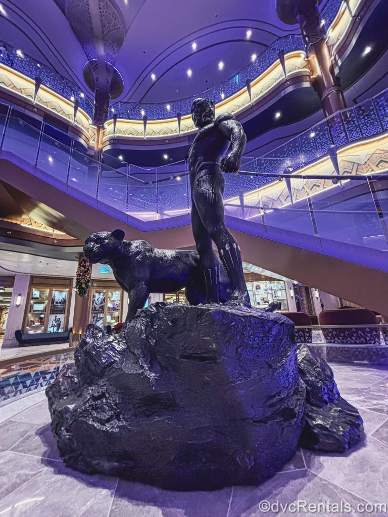 A statue of T’Challa, the Black Panther stands over the Grand Hall of the Disney Destiny under purple and yellow lighting.