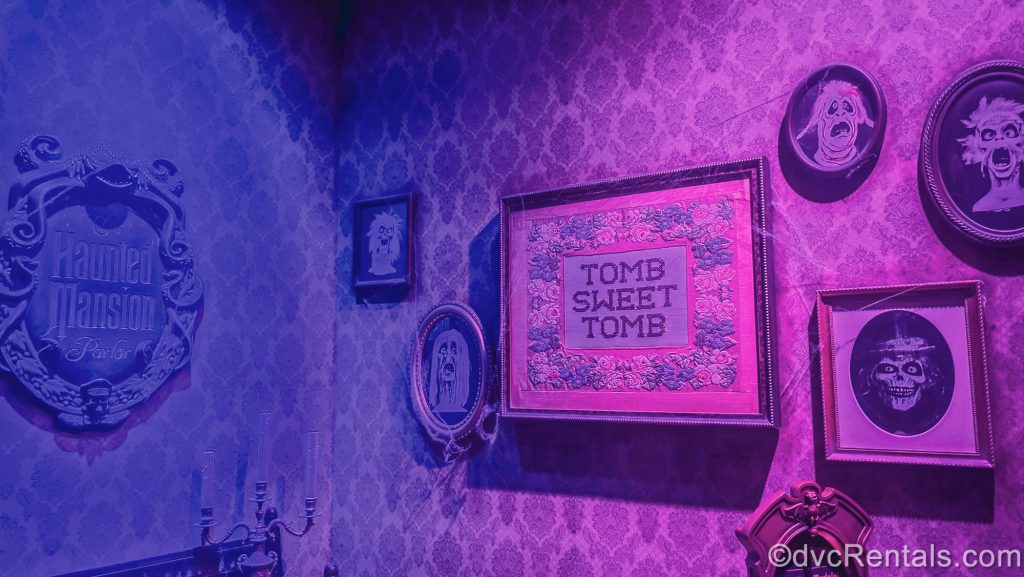 Themed signs reading “Haunted Mansion Parlor” and “Tomb Sweet Tomb” along with ghostly portraits hang in the entry hall to the Haunted Mansion Parlor under purple and pink lighting.