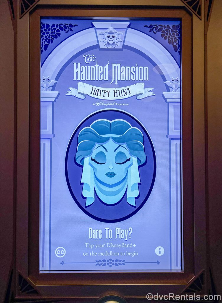 A poster-sized screen reading “The Haunted Mansion Happy Hunt, Dare to Play?” with a blue drawing of Madame Leota from the Disney Parks attraction is seen.