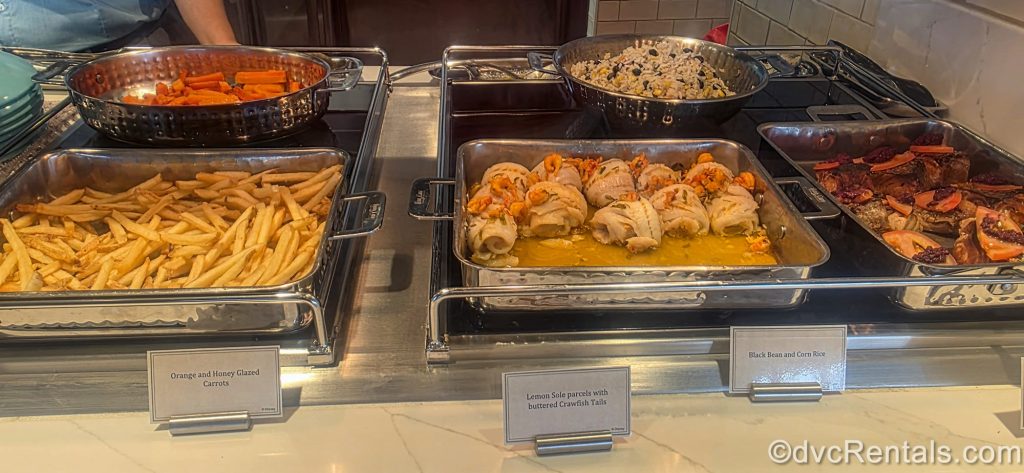 Silver trays displaying items such as seafood, chicken, and side dishes are seen on the Marceline Market buffet on the Disney Destiny.
