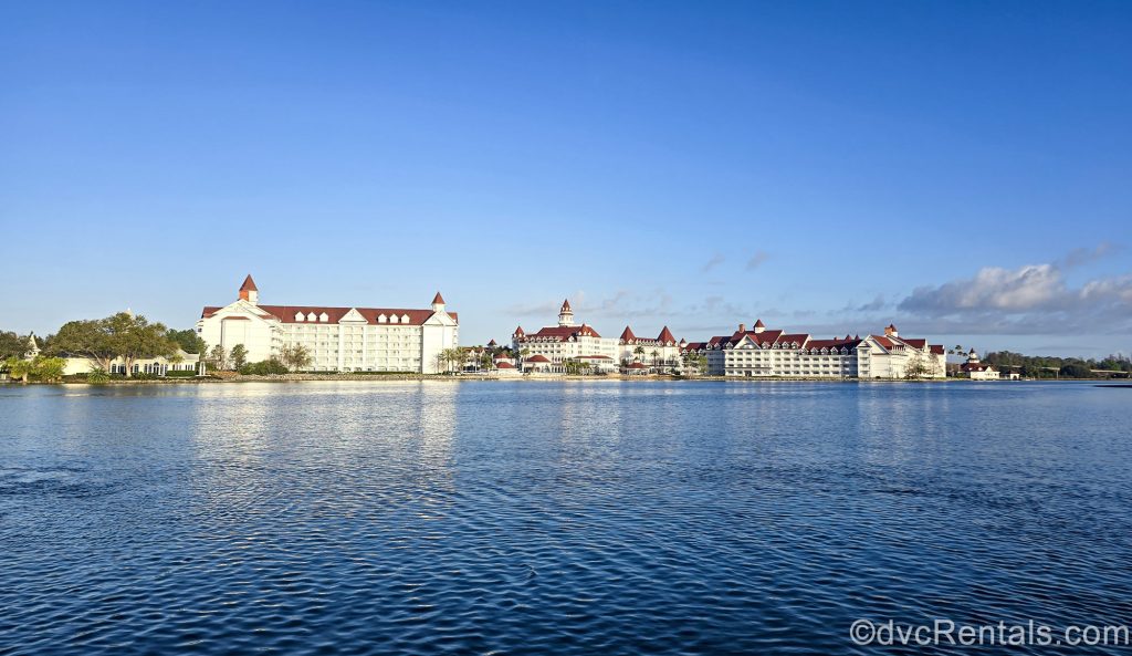 The white and red exterior of Disney’s Grand Floridian Resort & Spa is seen under a bright blue sky from across the water of the Seven Seas Lagoon.