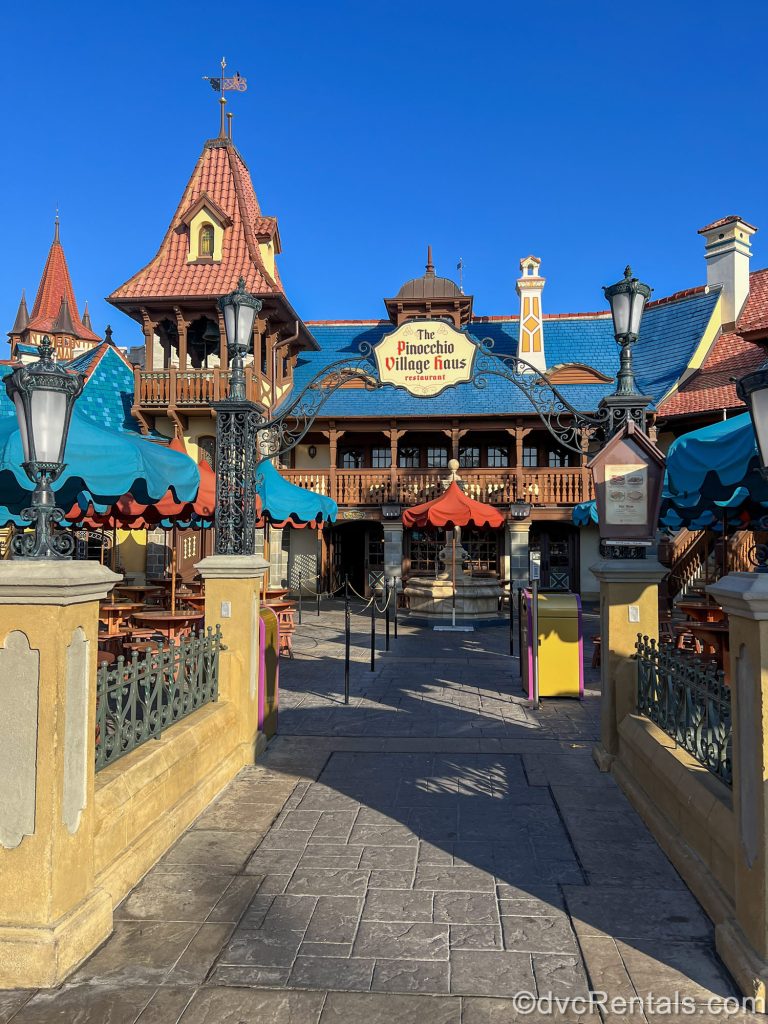 The exterior of Pinocchio Village Haus quick service restaurant and its outdoor seating areas are seen under a bright blue sky at Magic Kingdom.