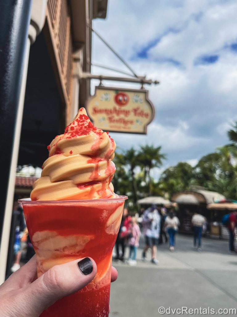 A hand holds up an “I Lava You” float made with orange Dole Whip at Sunshine Tree Terrace at Magic Kingdom. Red popping candies can be seen on top of the frozen treat.
