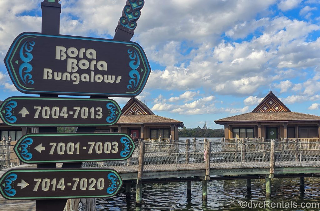 A dark wood and blue sign directs guests to the Bora Bora Bungalows at Disney’s Polynesian Villas & Bungalows. The Bungalows can be seen over the water behind the sign, under a blue sky, with Cinderella Castle visible in the distance.