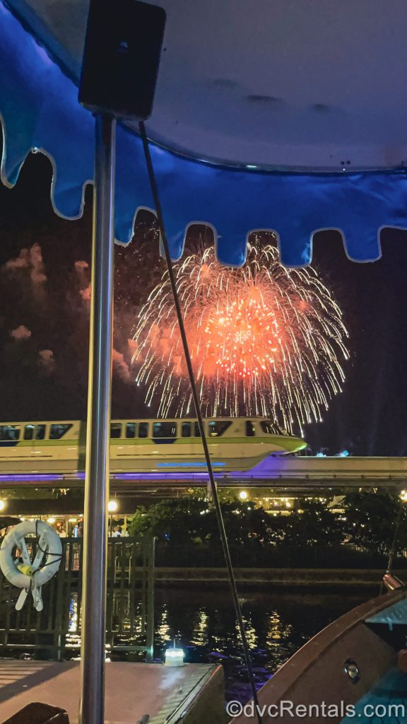 Red and white fireworks shine above the Magic Kingdom as seen from a water taxi boat as the monorail glides by under the night sky.