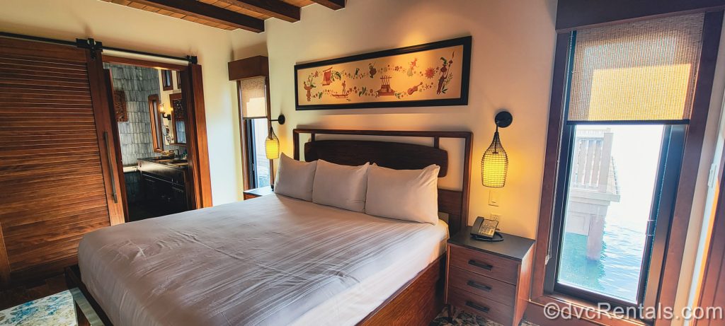 A bedroom in the Bungalows at Disney’s Polynesian Villas & Bungalows is seen. The room is decorated with dark woods and island-inspired accents, and the water of the Seven Seas Lagoon can be seen out the window.