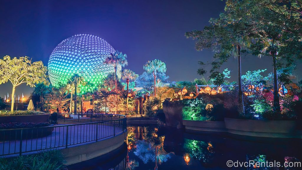 EPCOT’s grounds and Spaceship Earth icon are lit up in blue, green, and yellow lights under a night sky as the park prepares to close.
