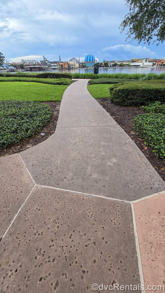A paved walking path leads from Disney’s Saratoga Springs Resort & Spa to Disney Springs under a cloudy sky.