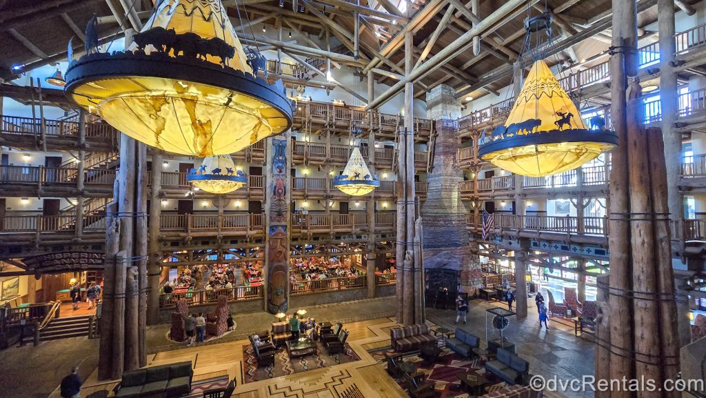 The grand, log cabin-style lobby of Disney’s Wilderness Lodge is seen. There are large wooden beams, Wilderness-inspired accents and art, seating spaces, and Whispering Canyon Café restaurant is also visible.