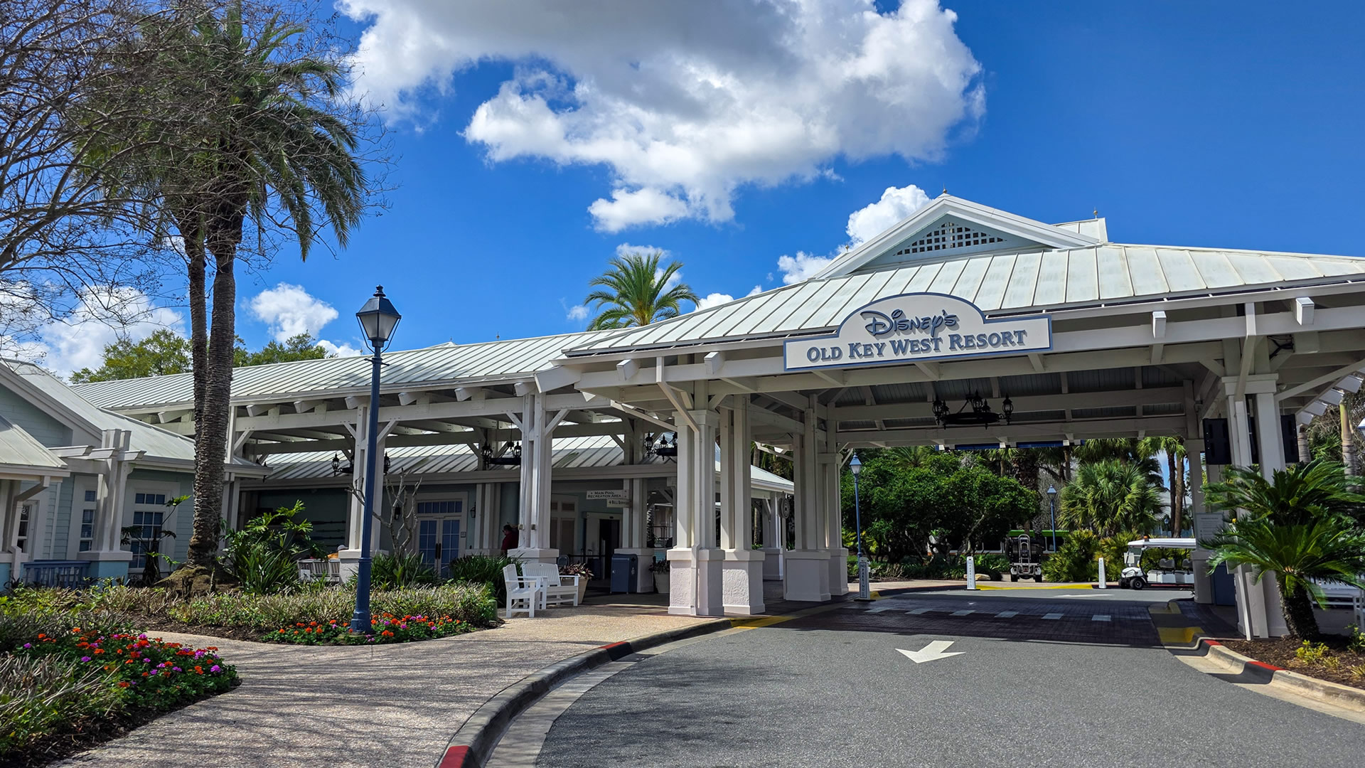 The white and blue porte-cochere of Disney’s Old Key West Resort is seen under a blue sky. Palm trees and colorful flower patches can be seen along the pedestrian walkways.