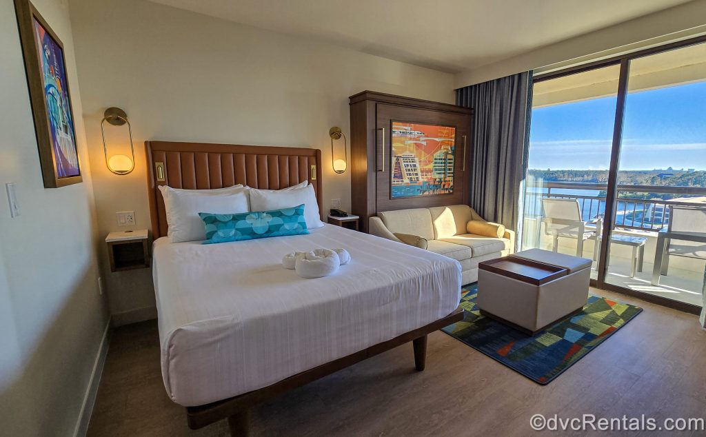 A Deluxe Studio at Bay Lake Tower at Disney’s Contemporary Resort. The room is decorated with neutral tones and bright Disney-themed art. A view of Bay Lake can be seen from the balcony door.