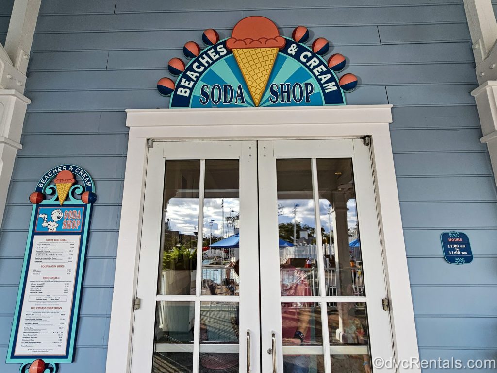 The entry door to Beaches & Cream Soda Shop is seen at Disney’s Beach Club Villas. Above the door is a blue sign with the restaurant’s name and it is decorated with coral-colored ice cream cones.