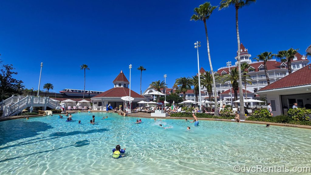 Guests enjoy the Beach Pool at Disney’s Grand Floridian Resort & Spa under a bright blue sky as the monorail glides by in the distance.