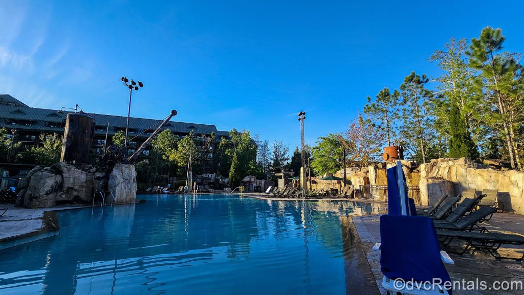 The Boulder Ridge Cove Pool is seen under a blue sky at Disney’s Wilderness Lodge. There are lounge chairs surrounding it and the resort’s exterior can be seen in the background.