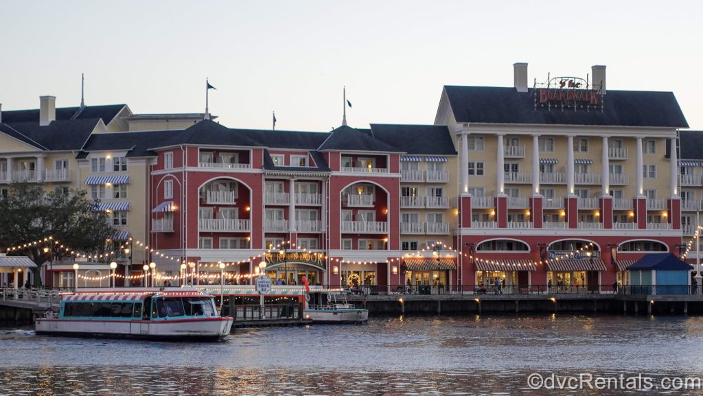 String lights shine outside the yellow and coral exterior buildings of Disney’s BoardWalk Villas at twilight.