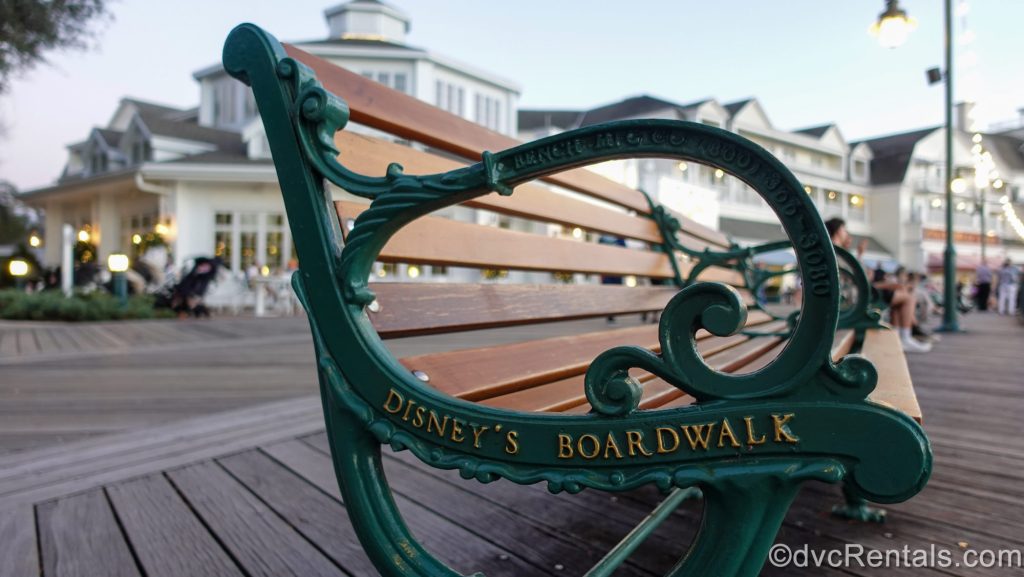 The words “Disney’s Boardwalk” are seen in gold script on the side of a wooden bench with green accents as the resort’s exterior is seen in the background.
