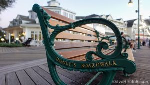 The words “Disney’s Boardwalk” are seen in gold script on the side of a wooden bench with green accents as the resort’s exterior is seen in the background.