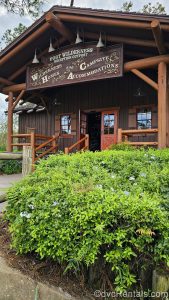 The log cabin-style exterior of the lobby of Disney’s Fort Wilderness Resort is seen under a cloudy sky. Above the doorway is a brown sign with white writing that reads “Fort Wilderness Reception Outpost. Wilderness Homes and Campsite Accommodations.”
